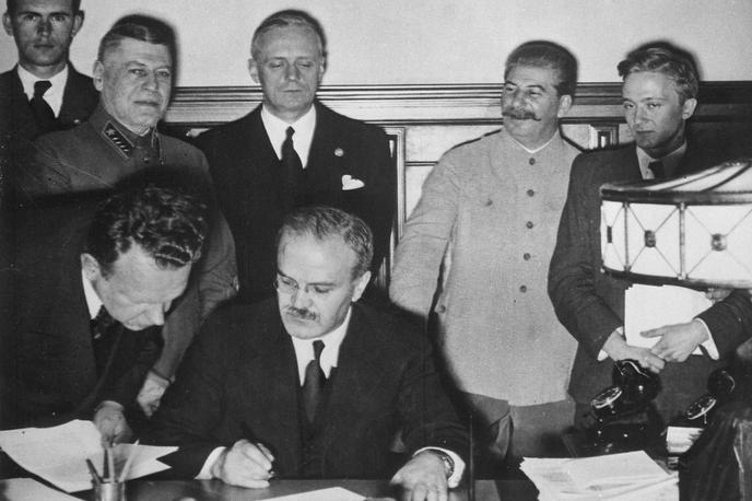 Podpisovanje pakta Ribbentrop-Molotov | Podpis pakta Ribbentrop-Molotov v Moskvi. Sovjetski zunanji minister Vjačeslav Molotov (z očali) sedi za mizo in podpisuje pakt. Za njim stoji nemški zunanji minister Joachim von Ribbentrop. Desno ob njem je Stalin, ki je več kot očitno zelo dobre volje. | Foto Wikimedia Commons